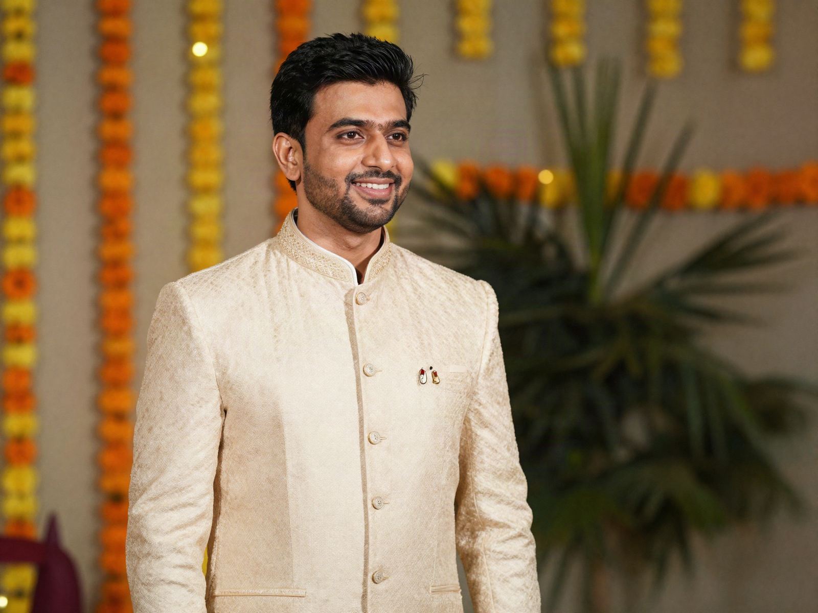 A man in a cream bandhgala at a wedding, marigold decorations behind him